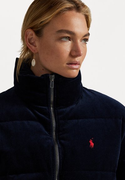 Donkerblauwe corduroy pufferjack met hoge kraag en frontale ritssluiting. Voorzien van een klein rood Polo-logo op de borst. Soepele textuur, gewatteerd ontwerp.