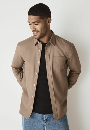 Brun skjorte med knapper og lomme, lavet af glat stof. Bæres over en sort skjorte, parret med lyseblå denimjeans.