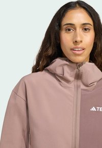 Veste légère de couleur rose-brun avec un col haut, une fermeture éclair intégrale et un logo sur la poitrine. Tissu lisse au toucher doux.