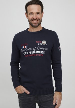 Marinblå strikket genser med tekstgrafikk i hvitt og rødt. Har ribbestrikkede mansjetter og nederkant, rund hals og et merke på venstre arm.