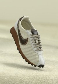 Bež umotani tenisice s smeškim kožnim Nike logom, svijetlo smeš tim đonom i uzorkovanim vezicama na mekanom teksturiranom bež površini.