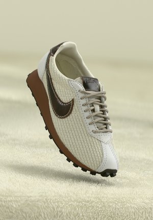 Beige woven sneaker with brown leather Nike swoosh, tan sole, and patterned laces on soft textured beige surface.