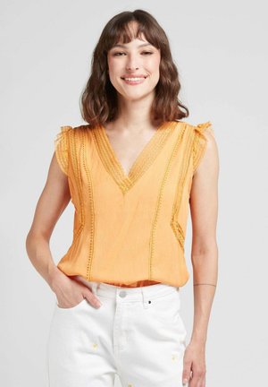 Blouse orange à col en V avec liseré de dentelle et manches volantes. Portée avec un pantalon blanc taille haute présentant des détails de broderie florale.