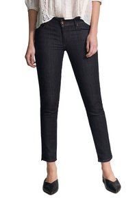 Femme portant un jean skinny foncé, un chemisier blanc avec des détails en dentelle et des chaussures slip-on noires à bout pointu, debout devant un fond blanc.