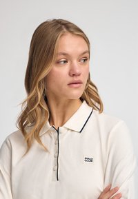 Polo shirt crema con accenti blu navy, caratterizzato da un colletto con bottoni e un piccolo logo "POLO CLUB" sul petto. Texture liscia e design casual.