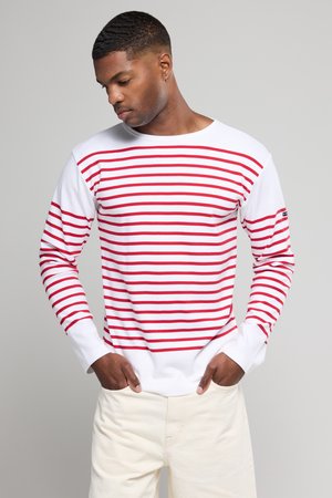 Chemise à manches longues blanche avec de larges rayures horizontales rouges, en tissu mélangé coton, encolure ronde et poignets ajustés. Détail de marque minimaliste.