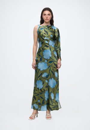 Femme portant une robe en satin vert à une épaule avec un grand imprimé floral bleu et des sandales blanches à talons ouvertes, debout devant un fond blanc.