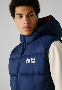 Chaleco acolchado azul marino con capucha, con cierre frontal de cremallera y logo "Red Bull RACING" en blanco. Material suave y brillante con diseño acolchado.