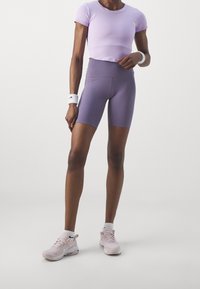 Woman wearing lavender crop top, high-waisted purple bike shorts, pink sneakers, and white wristbands against a plain background.