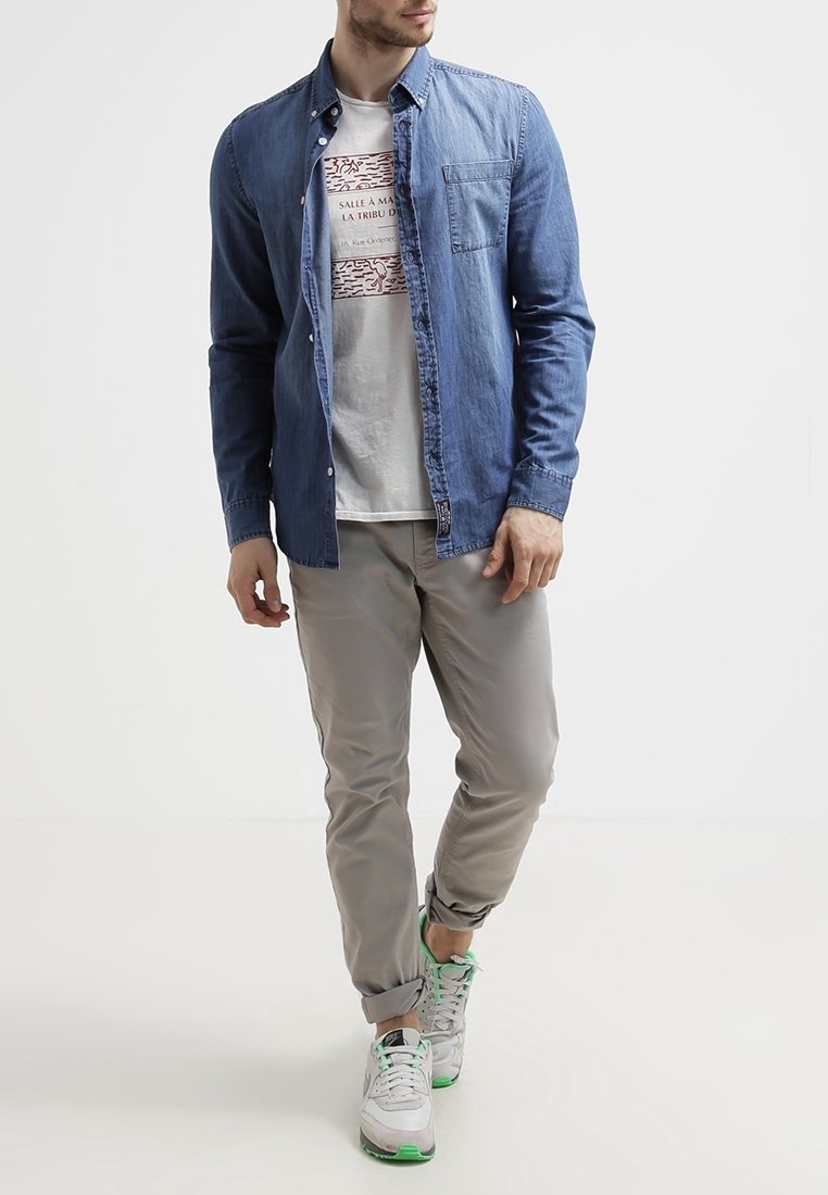 Camisa de mezclilla azul tipo button-up sobre una camiseta gráfica blanca, combinada con pantalones gris claro y zapatillas blancas con detalles en verde.