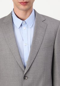 Giacca di suit grigia con texture, colletto a punte e un solo bottone. Sotto, una camicia azzurra con bottoni e colletto classico.