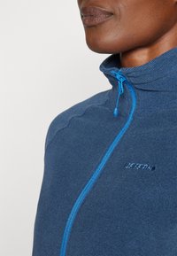 Blå zip-up jacka med ribbad textur och hög krage. Har en kontrasterande blå dragkedja och subtil logotypdetalj på bröstet.