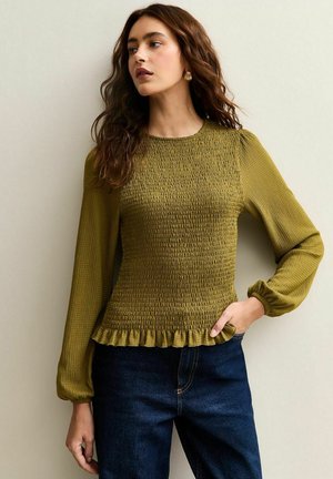 Blusa smock verde oliva con maniche lunghe a sbuffo e bordo arricciato, realizzata in tessuto strutturato. Abbinata a jeans in denim scuro.