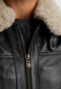 Črna usnjena bunda z bež ovčkom na ovratniku, zlatim zadrgo in strukturnim dizajnom, ki poudarja teksturirano površino in detajle šivanja.