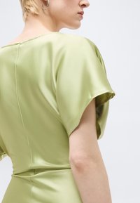 Άτομο με κοντά ξανθά μαλλιά που φοράει ανοιχτό πράσινο σατέν φόρεμα και ασημένιο κρίκο σκουλαρίκι, εμφανίζεται από πίσω και πλάγια.