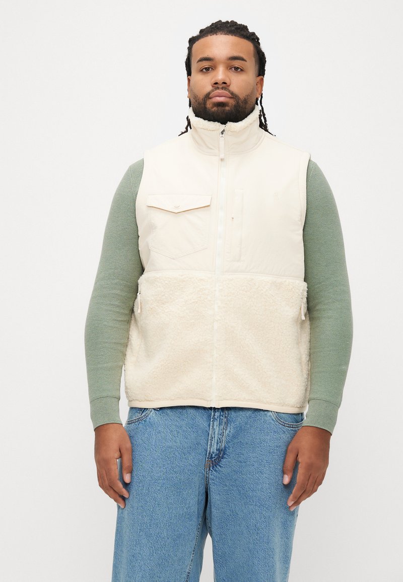 Colete bege sem mangas com parte inferior em pelo polar, fecho frontal com zip e bolso ao peito; usado sobre uma camisa verde de manga comprida. Jeans de ganga padrão.