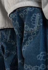 Denim jeans featuring a faded blue color with a repeating light blue chain pattern. The fabric has a textured finish and visible stitching details.