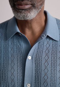 Jasnoniebieska dzianinowa koszulka polo z teksturowanym wzorem w diamenty, z szpiczastym kołnierzykiem i dwoma białymi guzikami przy dekolcie.