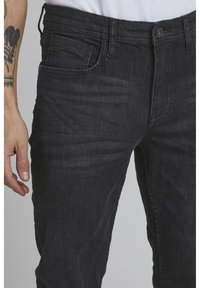Black denim jeans featuring a relaxed fit, front pockets, a zip fly, and a subtle texture. The waistband includes belt loops and a single button closure.
