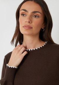 Brauner Strickpullover mit weißem gewelltem Saum am Hals und an den Ärmeln. Glatte Textur, runder Halsausschnitt und enganliegende Ärmel.