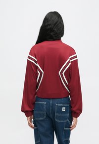 Kobieta z długimi ciemnymi włosami, ubrana w czerwoną kurtkę w białe i czarne paski oraz niebieskie dżinsowe spodnie cargo, zwrócona tyłem do aparatu.