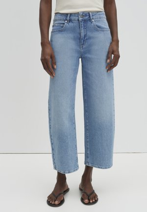 Person trägt hellblaue Jeans mit hohem Bund und geradem Bein sowie schwarze Flip-Flops, steht vor einem einfarbigen hellgrauen Hintergrund.