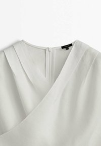 Vêtement blanc à col en V avec un design en tissu croisé et une fermeture éclair dans le dos visible en haut au centre.