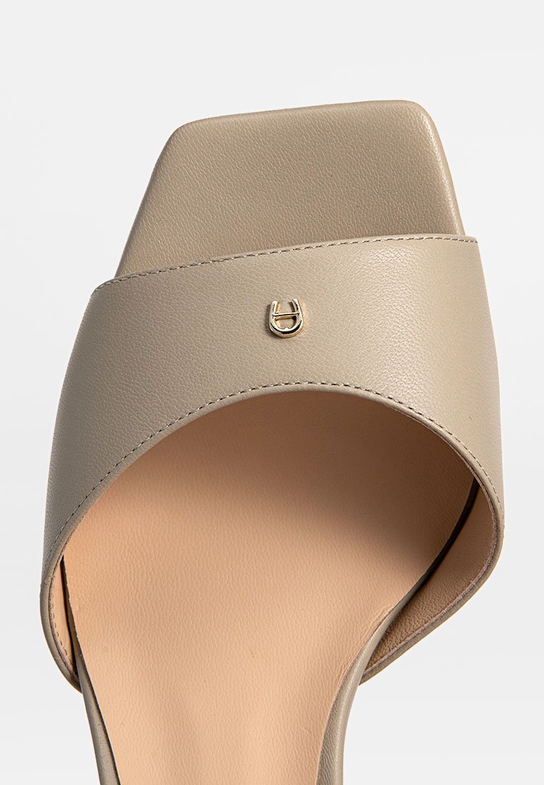 Sandale à talons en cuir beige avec un bout carré, dotée d'une large sangle et d'un accent logo doré. Texture lisse avec une semelle intérieure de couleur claire.