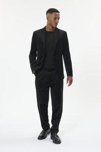 Mees, kes kannab musta sametist bleiserit, musta särki, lahtiseid musta sametist pükse ja musti nahkkingi, seisab käed taskutes.