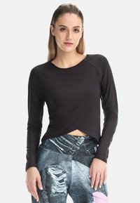 Ung kvinna med rakt blont hår bär en figurnära svart långärmad topp och slitna patchwork-jeans mot en vit bakgrund.