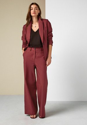 Frau in einem burgunderroten Blazer und weitgeschnittenen Hosen mit einem schwarzen V-Ausschnitt-Top, die mit den Händen in den Taschen vor einem beigen und weißen Hintergrund steht.