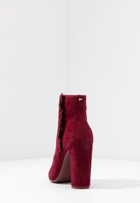 Stivale alla caviglia borgogna in suede, con tacco alto a blocco e un design arricciato lungo l'area della cerniera, arricchito da un piccolo accento dorato.