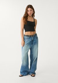 Top court noir associé à un jean large bleu clair et une ceinture noire avec des œillets en argent. Modèle posant sur un fond uni.