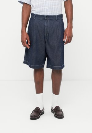 Uomo che indossa pantaloncini in denim scuro abbinati a calzini bianchi e mocassini in pelle marrone scuro su sfondo bianco.