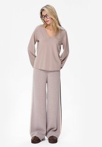 Lange mouwen, v-hals top in lichtbeige, gedragen met wijde, zachte grijze sweatpants met zijstrepen, nog eens aangevuld met donkerbruine sloffen.