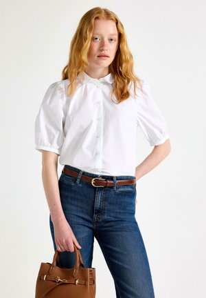 Kvinde med langt rødt hår iført en hvid kortærmet bluse, blå jeans, brun bælte og holder en brun håndtaske, står mod en enkel baggrund.