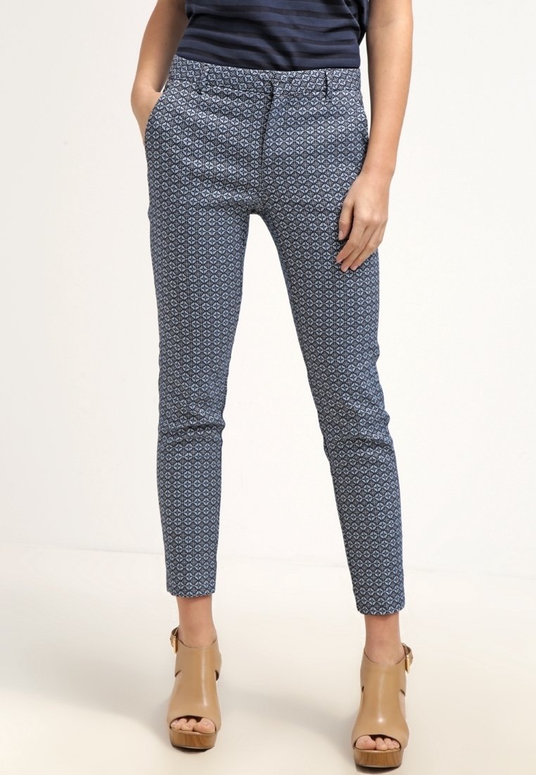 Femme portant un pantalon slim marine à motifs et des chaussures à talons ouverts couleur cuir clair, les mains dans les poches, sur un fond uni.