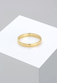 Simple polished gold band ring resting on a white geometric pedestal with a light gray background.