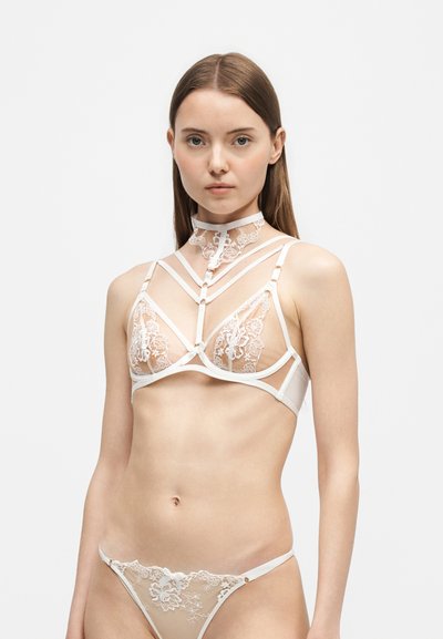 Junge Frau, die weiße florale Spitzenunterwäsche mit Riemchen-Details und Choker vor einfachem weißem Hintergrund trägt.