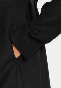 Cappotto nero in tessuto texturizzato, dotato di tasca laterale e maniche con risvolto, che presenta un design sartoriale e linee pulite.