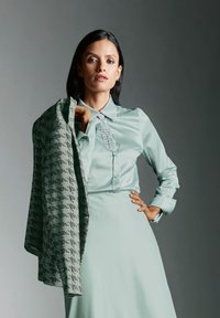Mintgroene blouse met een gestructureerde kraag en decoratieve knoopdetails, gecombineerd met een textuurgrijs-groene gestippelde jas.