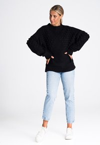 Czarny, dziany, oversized sweter z teksturowanym wykończeniem w formie bąbelków, z prążkowanymi mankietami i dołem, w połączeniu z jasnoniebieskimi, przetartymi dżinsami i białymi sneakersami.