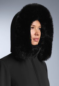 Crna parka s širokim lažnim krznenim kapuljačom, s glatkom teksturom tkanine, punim zatvaračem i minimalnim detaljima šivanja.