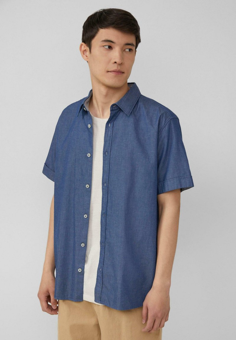 Camicia a maniche corte con bottoni di colore blu scuro, realizzata in cotone testurizzato, con colletto e orlo dritto, indossata sopra una T-shirt chiara.