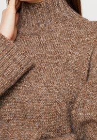 Un pull en tricot marron à col roulé avec des poignets côtelés et une texture montrant différentes nuances de marron dans le fil, créant une apparence douce.