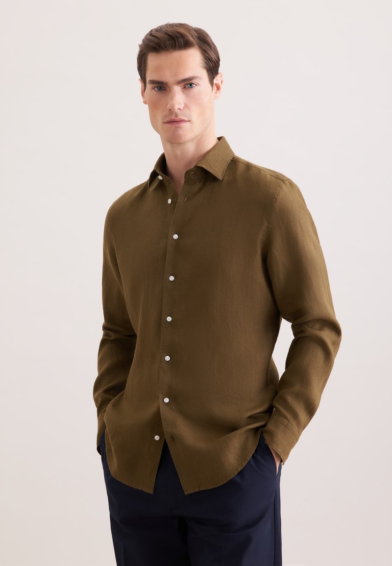 Homme portant une chemise à manches longues boutonnée vert olive avec col, debout les mains dans les poches devant un fond uni clair.