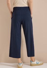 Personne portant un pantalon large bleu marine court avec une ceinture élastique et des baskets blanches avec des touches bleues, debout contre un mur beige.