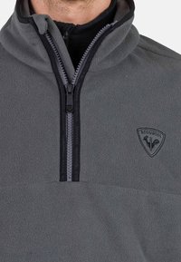 Pullover in pile grigio con zip nera e colletto interno, caratterizzato da una piccola patch a logo nera sul lato sinistro del petto e una texture liscia.