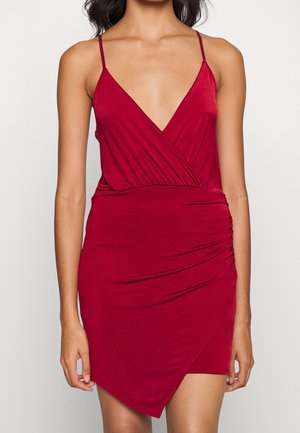 Femme portant une robe rouge sans manches avec de fines bretelles, un décolleté en V profond et un ourlet asymétrique avec des détails de tissu froncé.
