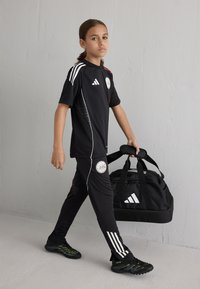 Tenue de sport noire comprenant un maillot avec des accents blancs et rouges, un pantalon noir avec des rayures sur le côté, et un sac de sport noir avec un logo blanc.